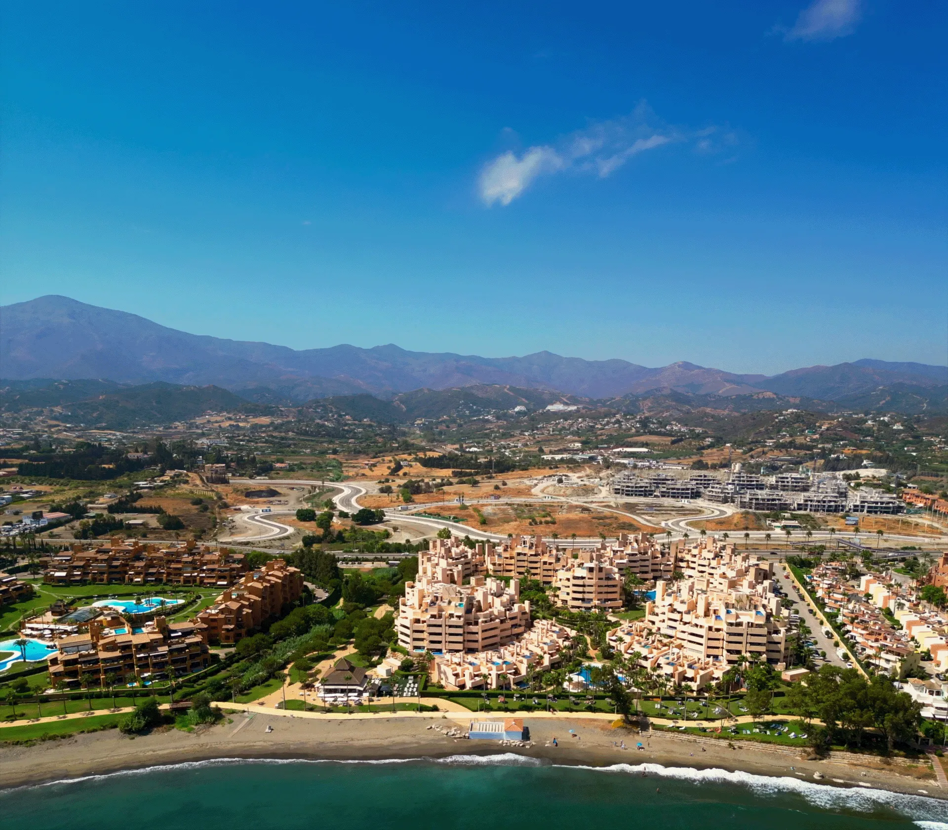 Coastal residential complex with pools, beachfront, roads, and distant mountains under clear blue sky.